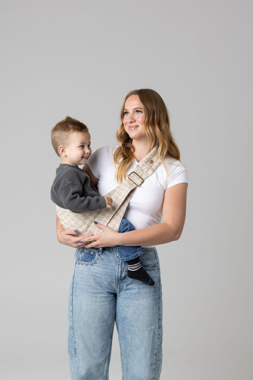 A brown-haired woman with blonde highlights standing face-forward against a white background looking off to the left of the frame. She wears light-wash jeans and a white t-shirt and has a toddler boy in the Mabē Monarch houndstooth-patterned toddler sling in beige and cream. It has a padded, quilted strap and a seat for the child to sit in, and a gold ladder-lock adjuster. The toddler is wearing a gray sweater and medium-wash jeans and black and gray socks and is looking to the right of the frame.