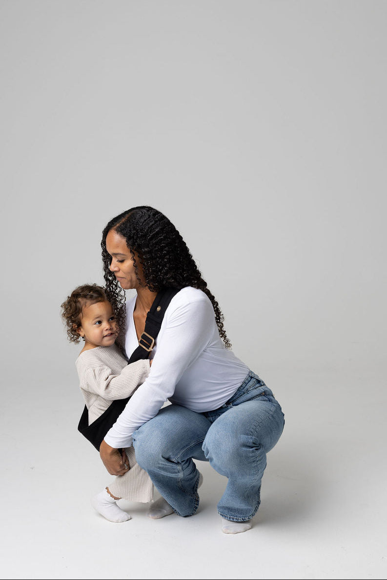 A tan-skinned woman with long, curly black hair kneels on the floor in a simple studio setting, gently supporting a toddler who stands close to her side, about to put the toddler up in a solid black Mabē toddler sling. She wears a white long‑sleeve shirt and blue jeans, while the child is dressed in cream colored coordinates with white socks.