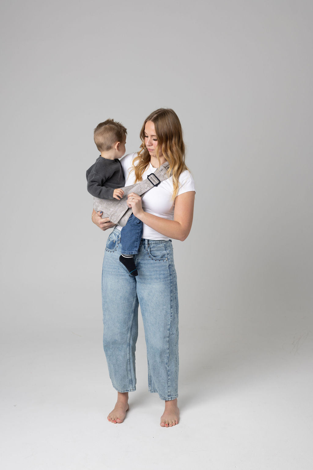 A brown-haired woman with blonde highlights standing face-forward against a white background wears light-wash jeans and a white t-shirt and has the toddler boy in a light gray Mabē Monarch toddler sling with Ashland Camo pattern, which she is looking down at. It has a padded, quilted strap and a seat for the child to sit in, and a black ladder-lock adjuster. The toddler is wearing a gray sweater and medium-wash jeans and black and gray socks.