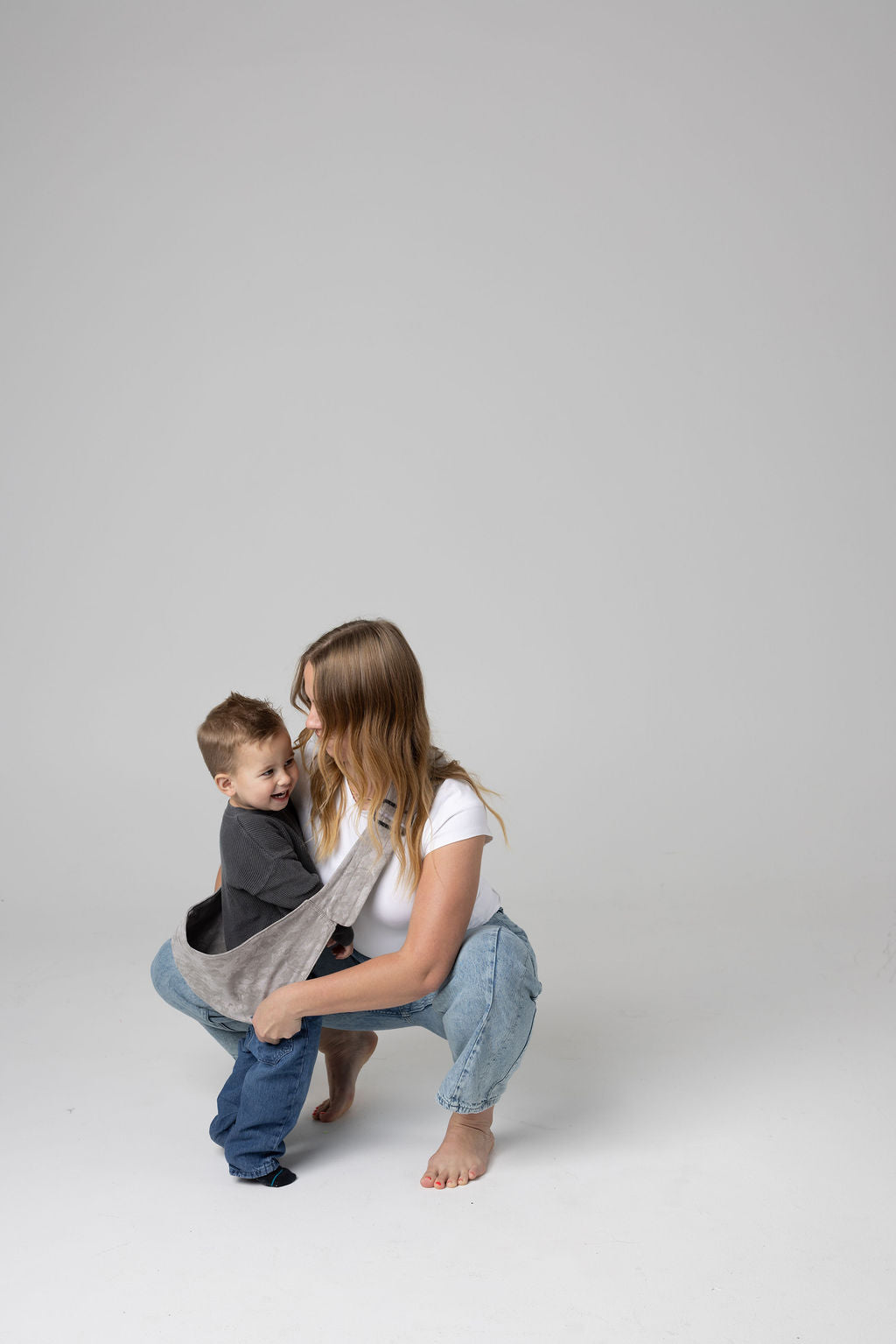 A brown-haired woman with blonde highlights crouched against a white background. She wears light-wash jeans and a white t-shirt and looks at a toddler boy being scooped in the light gray Mabē Monarch Ashland Camo toddler sling. It has a padded, quilted strap. The toddler is wearing a gray sweater and medium-wash jeans and black and gray socks and is looking to the right of the frame.