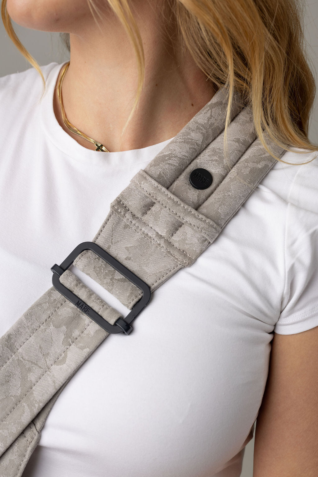 Close-up of the Mabē Monarch gray Ashland Camo toddler sling being worn by an unseen body with a white shirt and long reddish-blonde hair. It shows the padded, quilted strap with black ladder lock adjuster being worn cross-body.