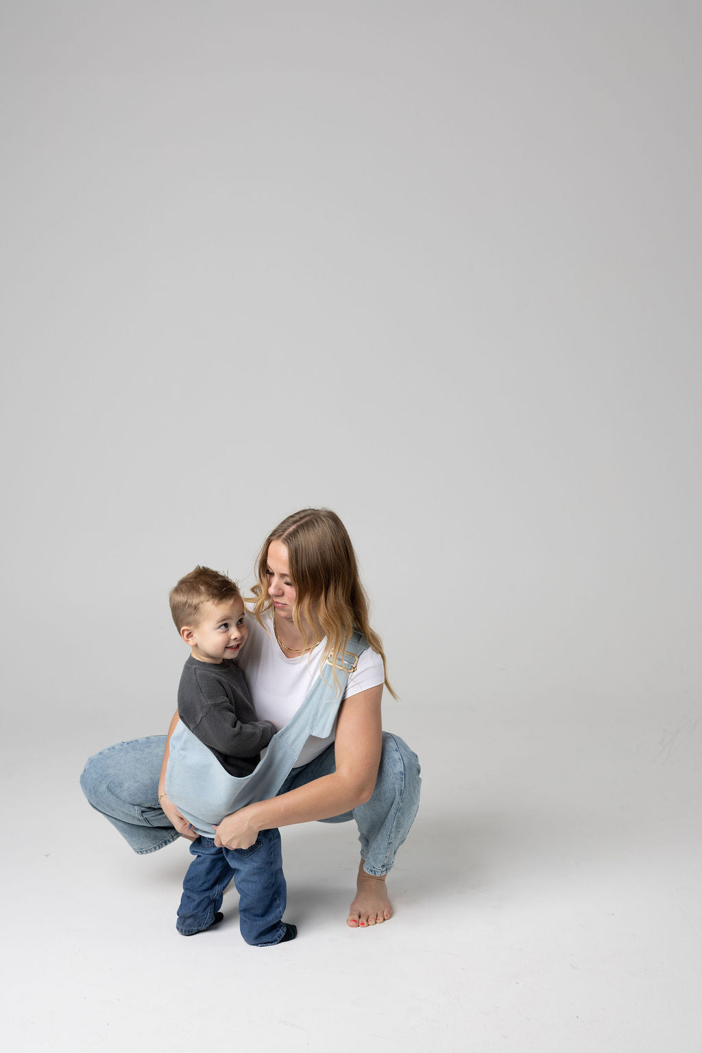A brown-haired woman with blonde highlights crouched against a white background. She wears light-wash jeans and a white t-shirt and looks at a toddler boy being scooped in the light blue Mabē Monarch Dream Wash Denim toddler sling. It has a padded, quilted strap. The toddler is wearing a gray sweater and medium-wash jeans and black and gray socks and is looking to the right of the frame.