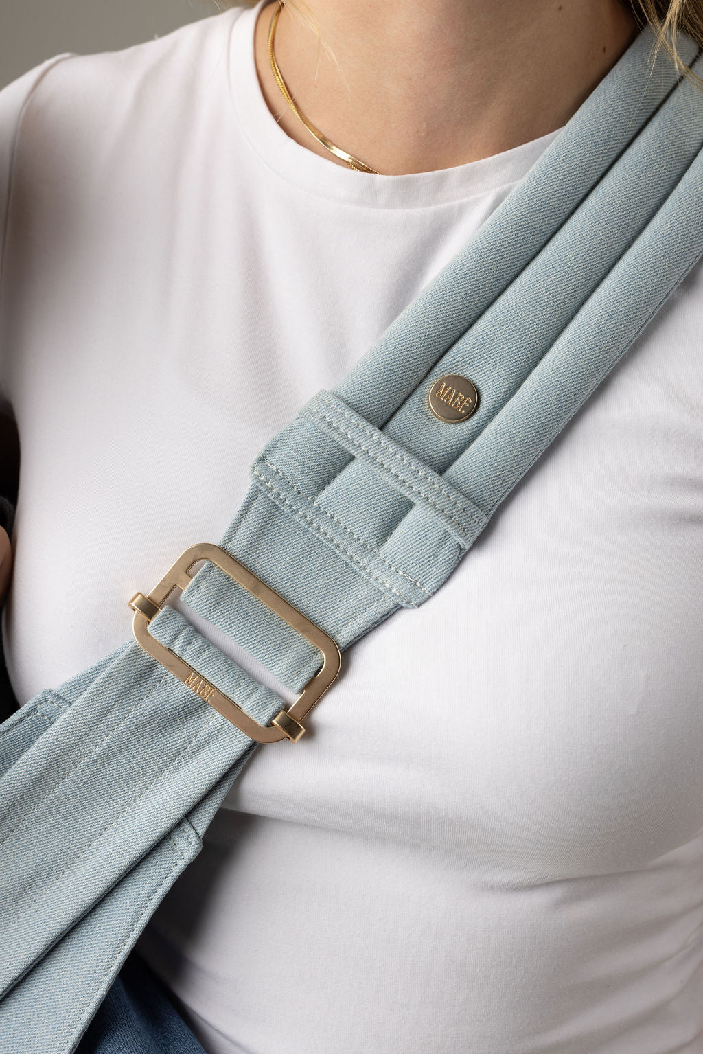 Close-up of the light blue Mabē Monarch Dream Wash Denim toddler sling being worn by an unseen body with a white shirt and long reddish-blonde hair. It shows the padded, quilted strap with gold ladder lock adjuster being worn cross-body.