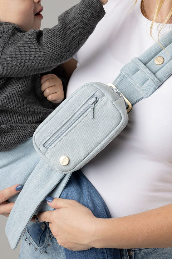 Close-up photo of an unseen body wearing light-wash jeans and a white t-shirt with a toddler in a light blue Mabē Monarch Dream Wash Denim toddler sling with matching accessory bag. The sling has a padded, quilted strap and a seat for the child to sit in, and a gold ladder-lock adjuster. The toddler is wearing a gray sweater and medium-wash jeans.