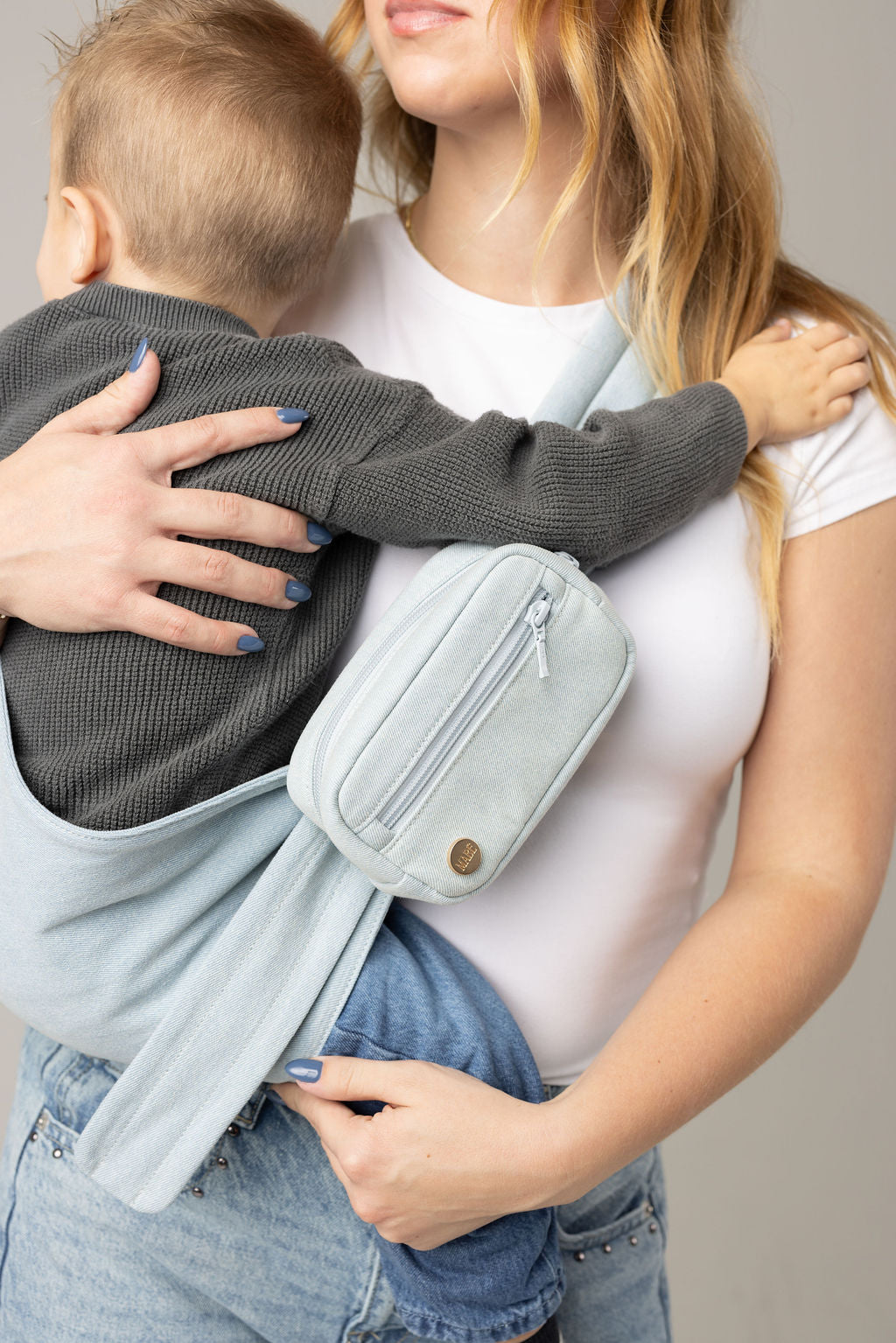A brown-haired woman with blonde highlights standing face-forward against a white background looking at her toddler in a sling carrier. She wears light-wash jeans and a white t-shirt and has a toddler boy in a light blue Mabē Monarch Dream Wash Denim toddler sling with matching accessory bag. The sling has a padded, quilted strap and a seat for the child to sit in, and a gold ladder-lock adjuster. The toddler is wearing a gray sweater and medium-wash jeans and black and gray socks.
