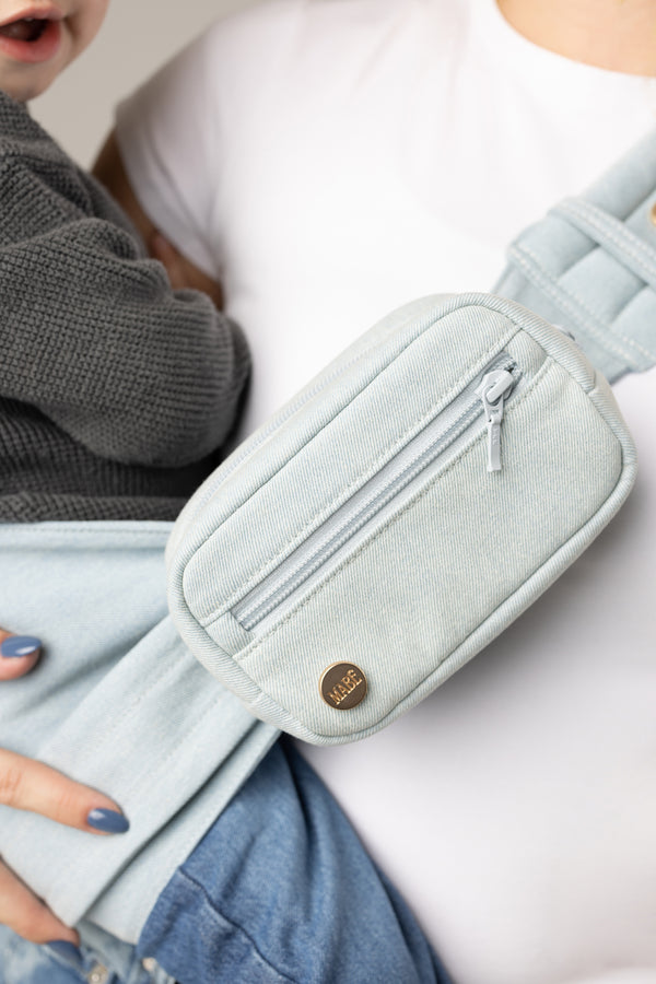 Light blue denim accessory bag worn by a person with a white shirt carrying a toddler in gray waffle shirt in the same denim Mabē toddler sling.