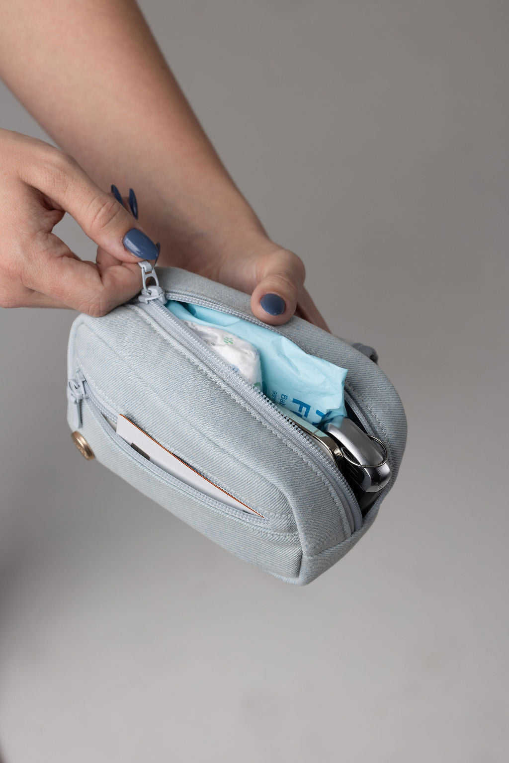 A light blue Dream Wash Denim accessory bag for Mabē slings and buckle carriers centered on a solid gray background. A hand with blue manicured nails unzips the main compartment. The small front compartment is already unzipped.