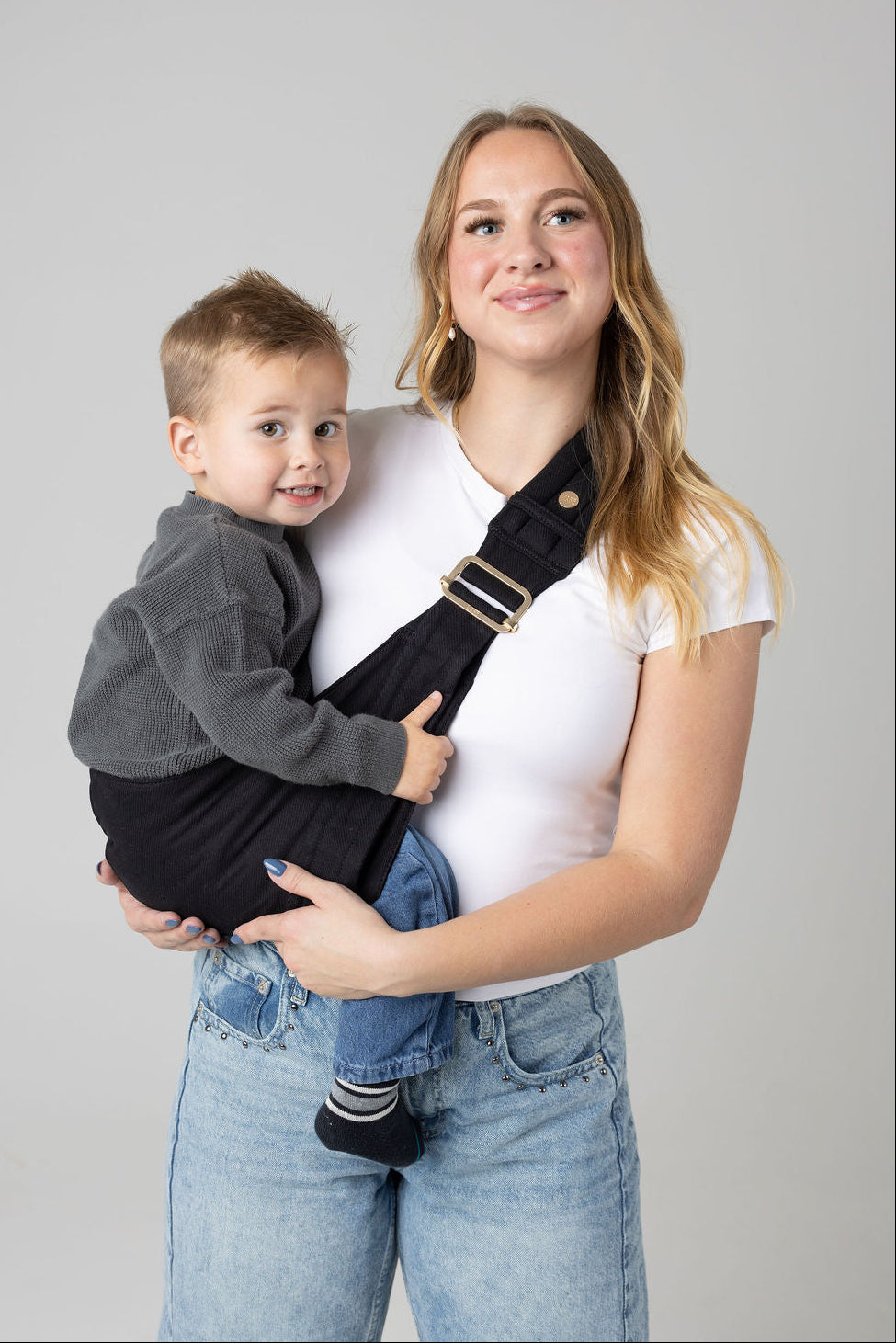 A photo of a brown-haired woman with blonde highlights from above the knees. She is standing straight forward against a white background with a toddler in a sling carrier. She wears light-wash jeans and a white t-shirt and has a toddler boy in a solid black Mabē Monarch toddler sling. It has a padded, quilted strap and a seat for the child to sit in, and a gold ladder-lock adjuster. The toddler is wearing a gray sweater and medium-wash jeans with black, gray, and white striped socks.
