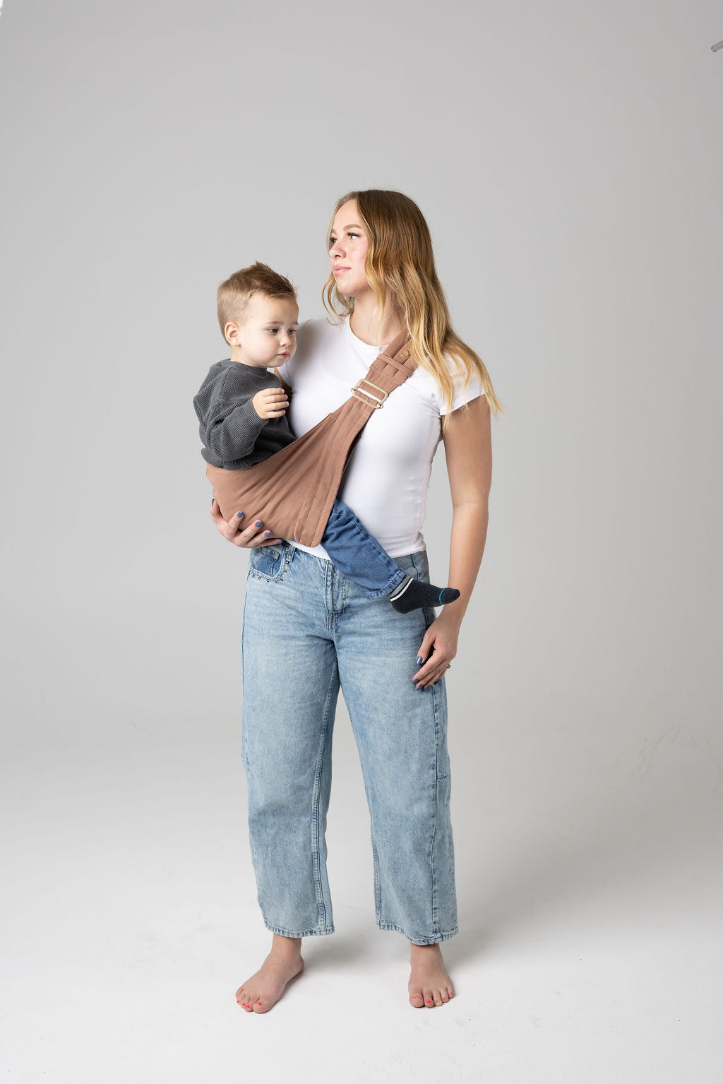 A brown-haired woman with blonde highlights standing face-forward against a white background looking at her toddler in a sling carrier. She wears light-wash jeans and a white t-shirt and has a toddler boy in a black and white checkered Mabē Monarch toddler sling. It has a padded, quilted strap and a seat for the child to sit in, and a gold ladder-lock adjuster. The toddler is wearing a gray sweater and medium-wash jeans and black and gray socks.