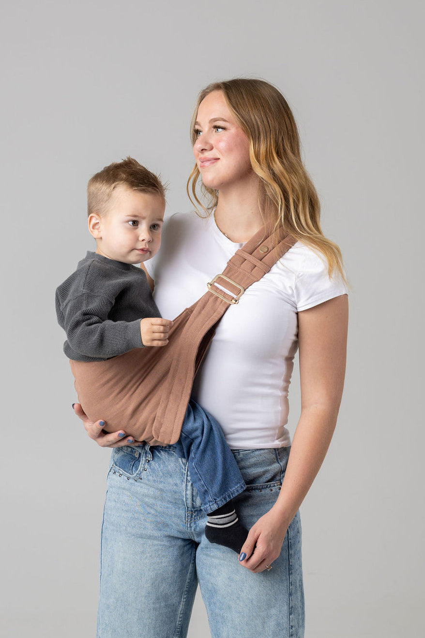 A brown-haired woman with blonde highlights standing face-forward against a white background looking at her toddler in a sling carrier. She wears light-wash jeans and a white t-shirt and has a toddler boy in a solid mocha-colored Mabē Monarch toddler sling. It has a padded, quilted strap and a seat for the child to sit in, and a gold ladder-lock adjuster. The toddler is wearing a gray sweater and medium-wash jeans and black and gray socks.