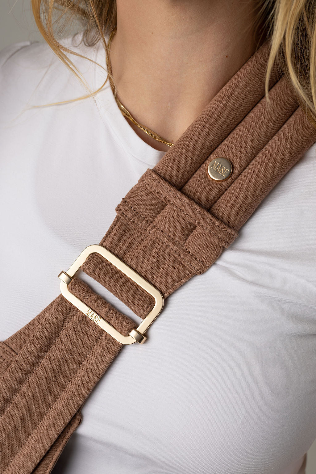 Close-up of the Mabē Monarch solid mocha-colored toddler sling being worn by an unseen body with a white shirt and long reddish-blonde hair. It shows the padded, quilted strap with gold ladder lock adjuster being worn cross-body.