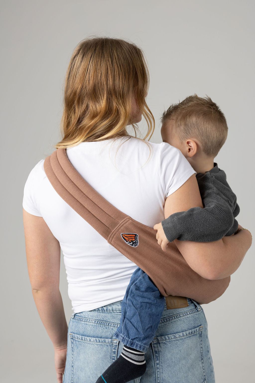 A light-skinned woman facing away from the viewer. She has long, reddish brown and blonde curled hair and is holding a tan-skinned toddler with light brown hair in a solid, mocha-colored Mabē toddler sling. The woman wears a long-sleeved white shirt and mid-wash jeans, and the toddler wears a gray sweater and jeans with black and gray socks.