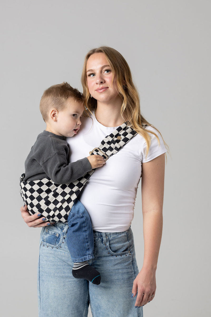 A brown-haired woman with blonde highlights standing face-forward against a white background looking at her toddler in a sling carrier. She wears light-wash jeans and a white t-shirt and has a toddler boy in a black and white checkered Mabē Monarch toddler sling. It has a padded, quilted strap and a seat for the child to sit in, and a gold ladder-lock adjuster. The toddler is wearing a gray sweater and medium-wash jeans and black and gray socks and cuddles the woman's shoulder.