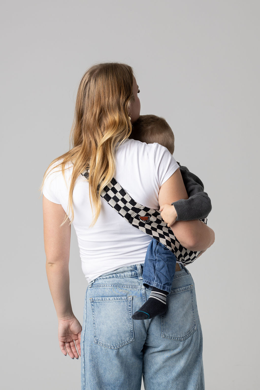 A light-skinned woman facing away from the viewer. She has long, reddish brown and blonde curled hair and is holding a tan-skinned toddler with light brown hair in a black and white checkered Mabē toddler sling. The woman wears a long-sleeved white shirt and mid-wash jeans, and the toddler wears a gray sweater and jeans with black and gray socks.