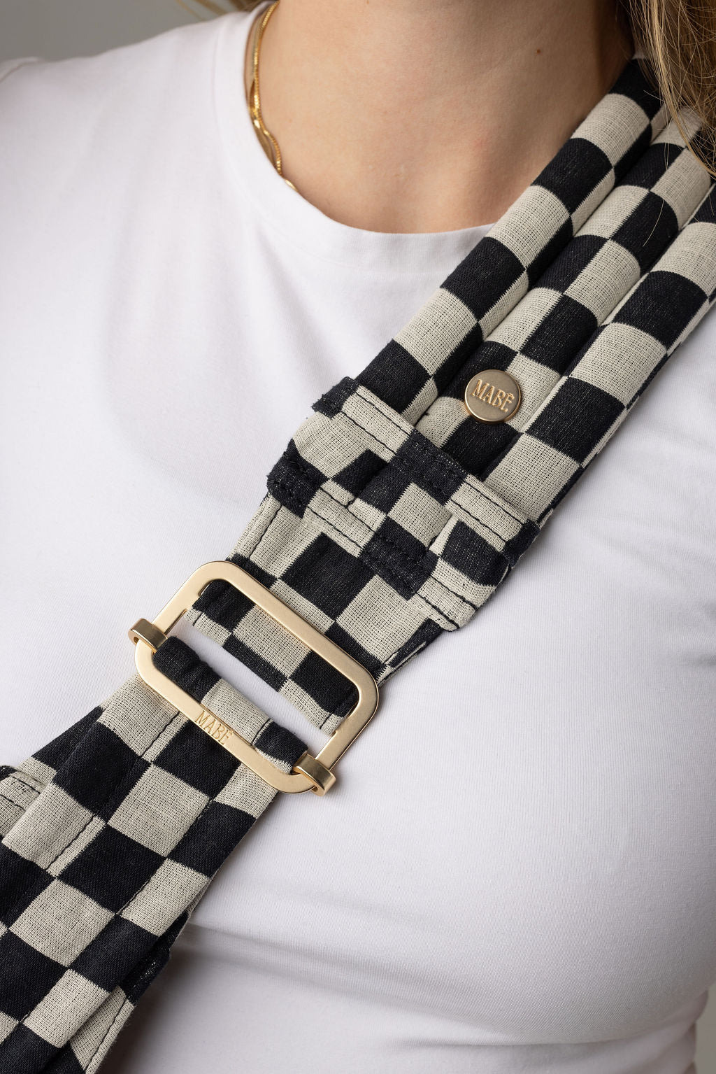 Close-up of the black and white checkered Mabē Monarch toddler sling being worn by an unseen body with a white shirt and long reddish-blonde hair. It shows the padded, quilted strap with gold ladder lock adjuster being worn cross-body.