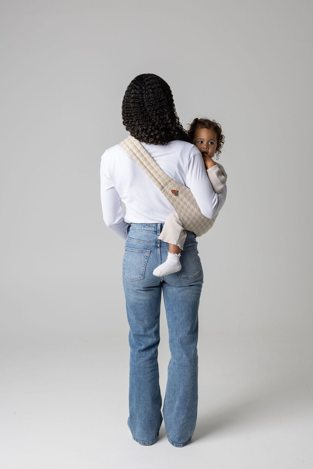 A brown-skinned woman facing away from the viewer. She has long, black, curly hair and is holding a tan-skinned toddler with brown, curly hair in a houndstooth patterned beige Mabē toddler sling. The woman wears a long-sleeved white shirt and mid-wash jeans, and the toddler wears cream-colored knitted coordinating long-sleeved top and bottom and white socks. They stand against a white background.