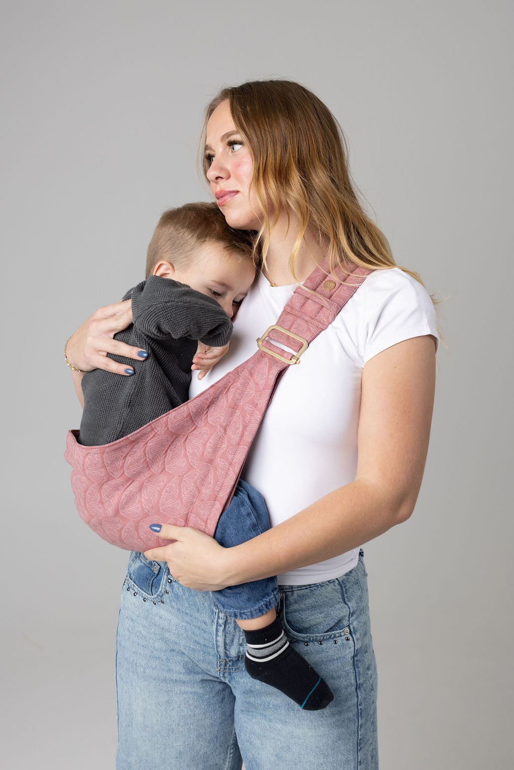 A brown-haired woman with blonde highlights standing face-forward against a white background looking at her toddler in a sling carrier. She wears light-wash jeans and a white t-shirt and has a toddler boy in a pink Mabē Monarch toddler sling with butterfly wing pattern. It has a padded, quilted strap and a seat for the child to sit in, and a gold ladder-lock adjuster. The toddler is wearing a gray sweater and medium-wash jeans and black and gray socks.