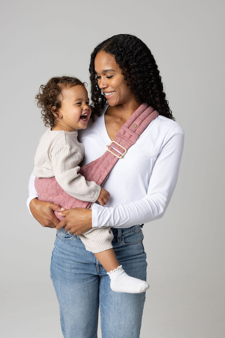 A tan-skinned woman with long curly black hair stands against a white backdrop, holding a laughing, tan-skinned, curly-haired toddler on her hip using a soft, quilted pink Mabē hip sling with patterned butterfly wings. It crosses her shoulder and fastens with a gold buckle. She wears a white long‑sleeve shirt and blue jeans, while the toddler is dressed in a light beige knit outfit with one small white sock showing. The child leans comfortably against her, creating a warm, close, and gentle scene.