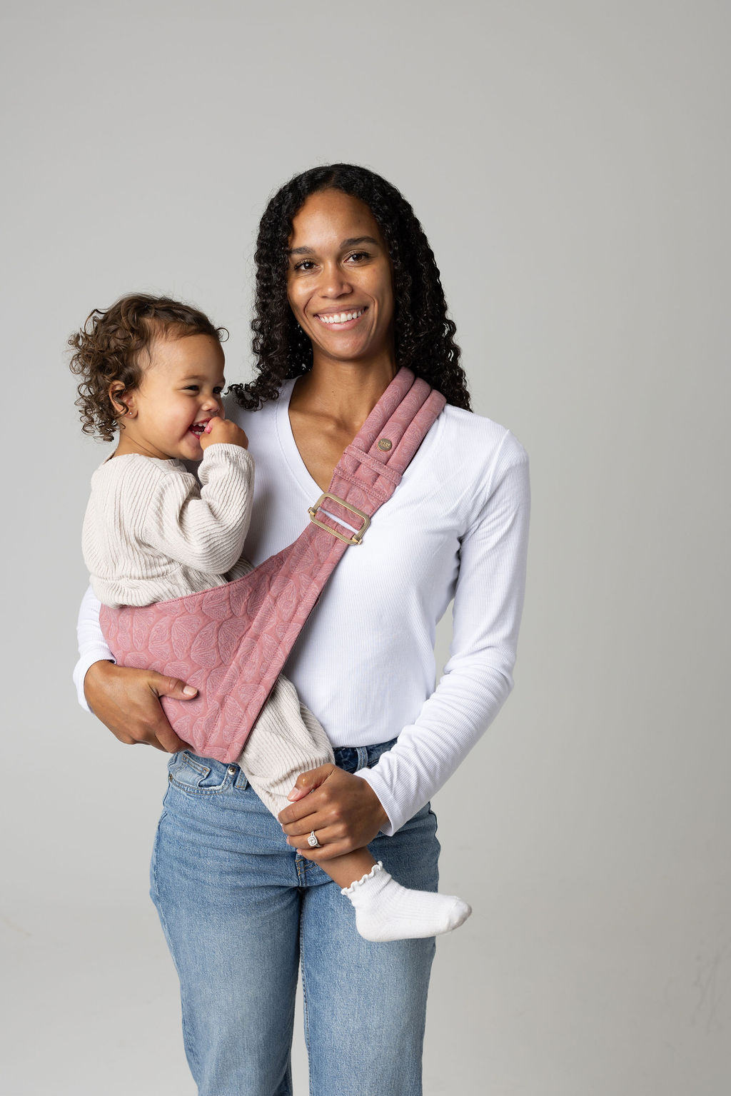 A brown-skinned woman with black curly long hair standing face-forward against a white background looking at her toddler in a sling carrier. She wears light-wash jeans and a white t-shirt and has a toddler  in a pink, butterfly-wing-patterned Mabē Monarch toddler sling. It has a padded, quilted strap and a seat for the child to sit in, and a gold ladder-lock adjuster. The toddler is wearing coordinating cream sweater and pants and white socks and is looking to the right of the frame.