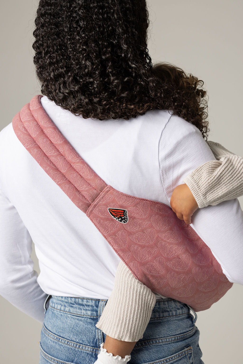 A woman with long curly black hair facing away from the viewer. She is holding a tan-skinned toddler with brown hair in a pink Mabē toddler sling patterned with butterfly wings. The woman wears a long-sleeved white shirt and mid-wash jeans, and the toddler wears cream knitted coordinates and white lettuce trim socks.
