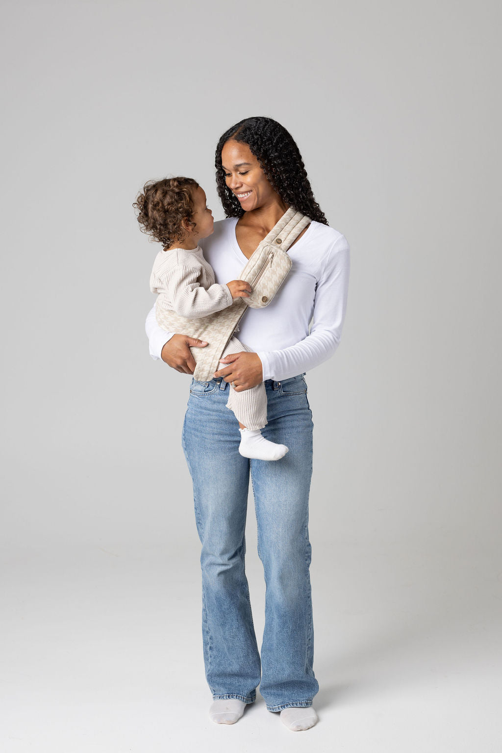 A brown-skinned woman with long, black, curly hair holding a tan-skinned toddler with brown, curly hair in a houndstooth patterned beige Mabē toddler sling with matching accessory bag. The woman wears a long-sleeved white shirt and mid-wash jeans, and the toddler wears cream-colored knitted coordinating long-sleeved top and bottom and white socks. They stand against a white background.
