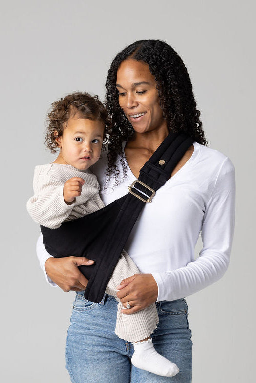 A brown-skinned woman with black curly long hair standing face-forward against a white background looking at her toddler in a sling carrier. She wears light-wash jeans and a white t-shirt and has a toddler  in a solid black Mabē Monarch toddler sling. It has a padded, quilted strap and a seat for the child to sit in, and a gold ladder-lock adjuster. The toddler is wearing coordinating cream sweater and pants and white socks and is looking to the left of the frame.