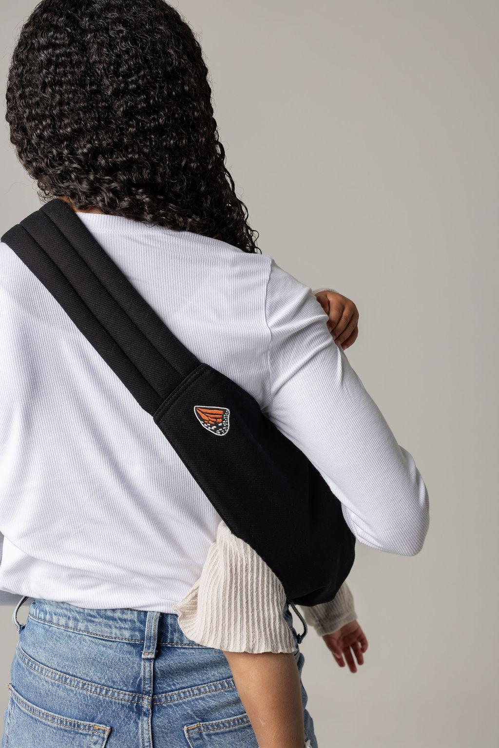 A brown-skinned woman with curly black hair facing away from the viewer shown from the hips up is holding a toddler hair in a solid black Mabē toddler sling. The woman wears a long-sleeved white shirt and mid-wash jeans, and the toddler wears cream-colored knit coordinates.