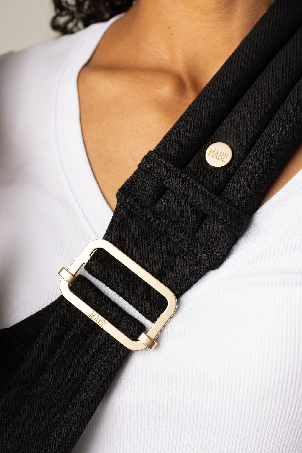Close-up of the Mabē Monarch solid black Ebony toddler sling being worn by an unseen body with a white shirt and brown skin. It shows the padded, quilted strap with gold ladder lock adjuster being worn cross-body.