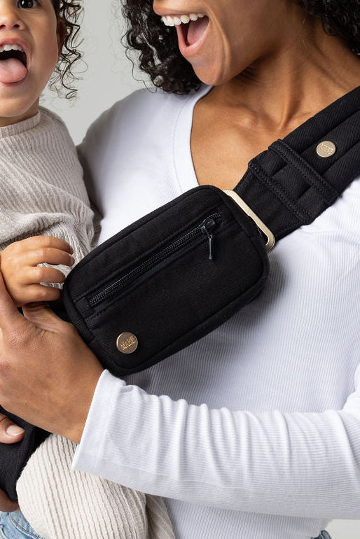 A closed-in photo of a tan-skinned woman with curly black hair standing face-forward against a white background. She wears light-wash jeans and a white t-shirt and has a toddler in a black Mabē Monarch  toddler sling with matching black accessory bag, which the toddler is unzipping. The sling has a padded, quilted strap and a seat for the child to sit in, and a black ladder-lock adjuster. The toddler is wearing cream knitted coordinates.