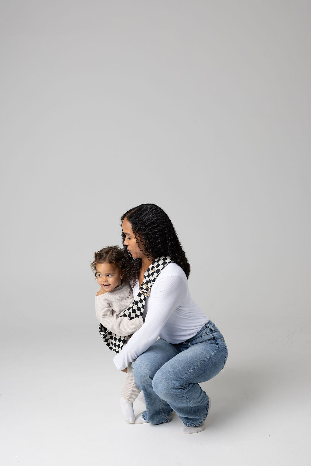 A black-haired woman with tan skin crouched against a white background. She wears light-wash jeans and a white t-shirt and looks at a toddler girl being scooped into the black and white checkered Mabē Monarch toddler sling. It has a padded, quilted strap. The toddler is wearing cream coordinates and white socks.