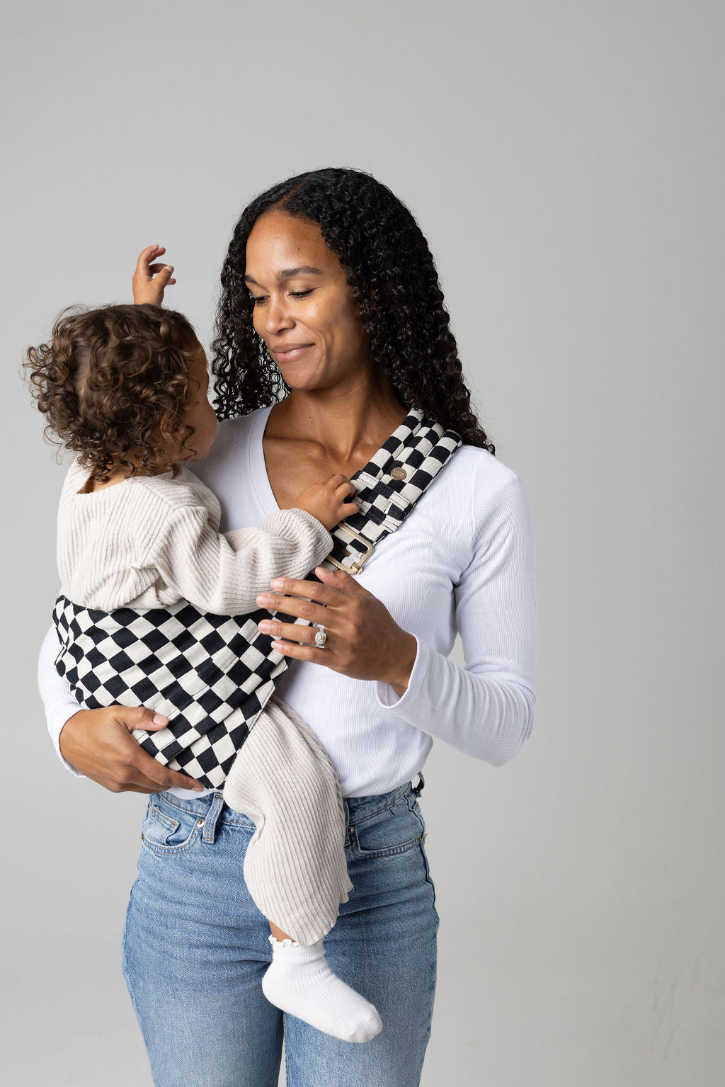 A brown-skinned woman with black curly long hair standing face-forward against a white background looking at her toddler in a sling carrier. She wears light-wash jeans and a white t-shirt and has a toddler boy in a black and white checkered Mabē Monarch toddler sling. It has a padded, quilted strap and a seat for the child to sit in, and a gold ladder-lock adjuster. The toddler is wearing coordinating cream sweater and pants and white socks and reaches into the woman's face.
