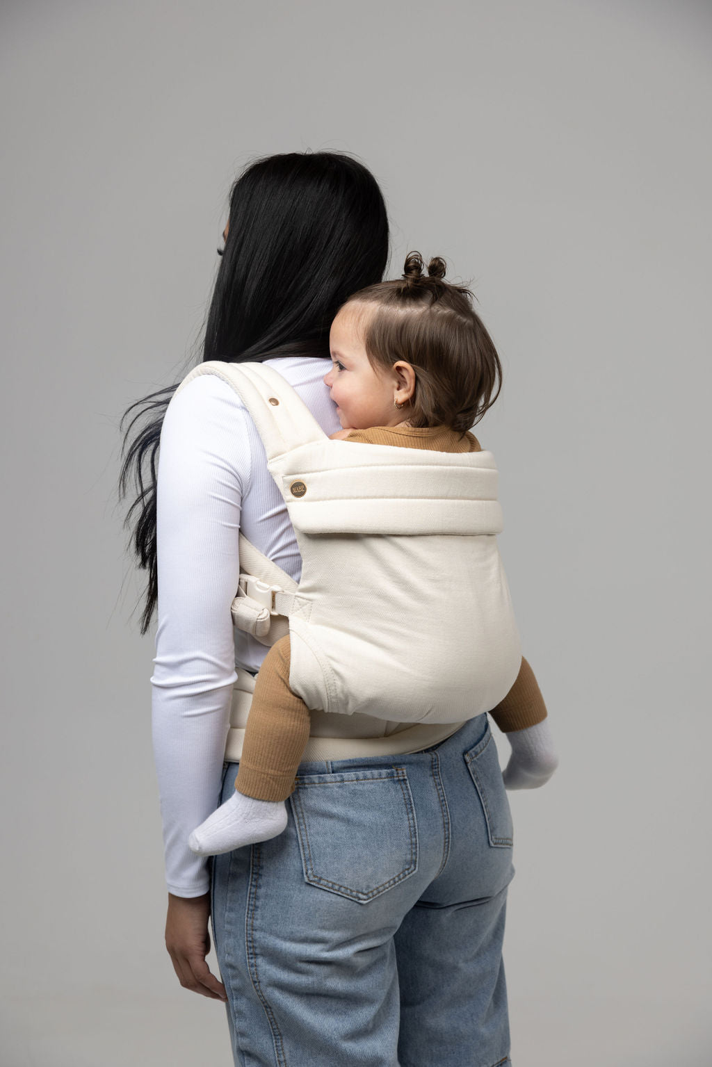 A person whose only visible features aside from their clothed body is long, straight, black hair and tan-skinned hand faces away from the viewer. They are wearing a white long-sleeved shirt and light-wash jeans and a toddler with short dark straight hair on their back in the Mabē Monarch Buckle Carrier in Ivory, which is solid warm off-white. The toddler wears tan pants. The background is solid white.
