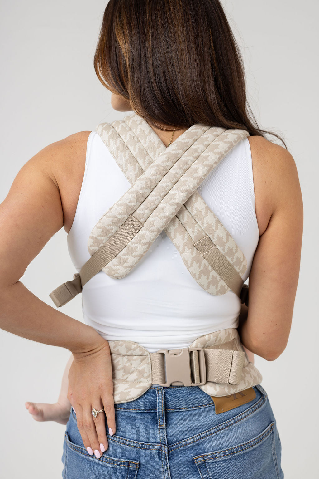 An unseen person with long brown hair with highlights and tanned skin is shown from behind. They wear a white tank top and the Mabē Monarch Buckle Carrier in Houndstooth, which is cream and beige. Only the carrier's straps crossed across the back and the waistband are visible. The top part of the person's medium wash jeans are visible but the picture is cropped mid-back-pocket and below.