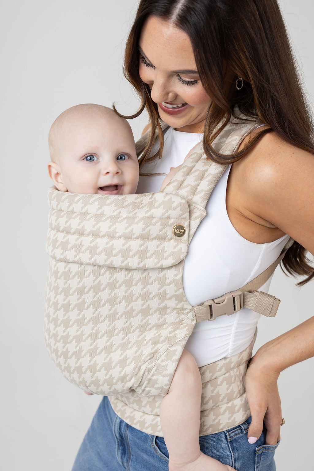 A woman with long brown hair with highlights and tanned skin wears a white tank top and the Mabē Monarch Buckle Carrier in Houndstooth, which is cream and beige. Inside is a light-skinned baby with blue eyes. She wears medium wash jeans and looks down at the baby with a big smile. The baby is looking at the viewer with a smile. His legs are bare.