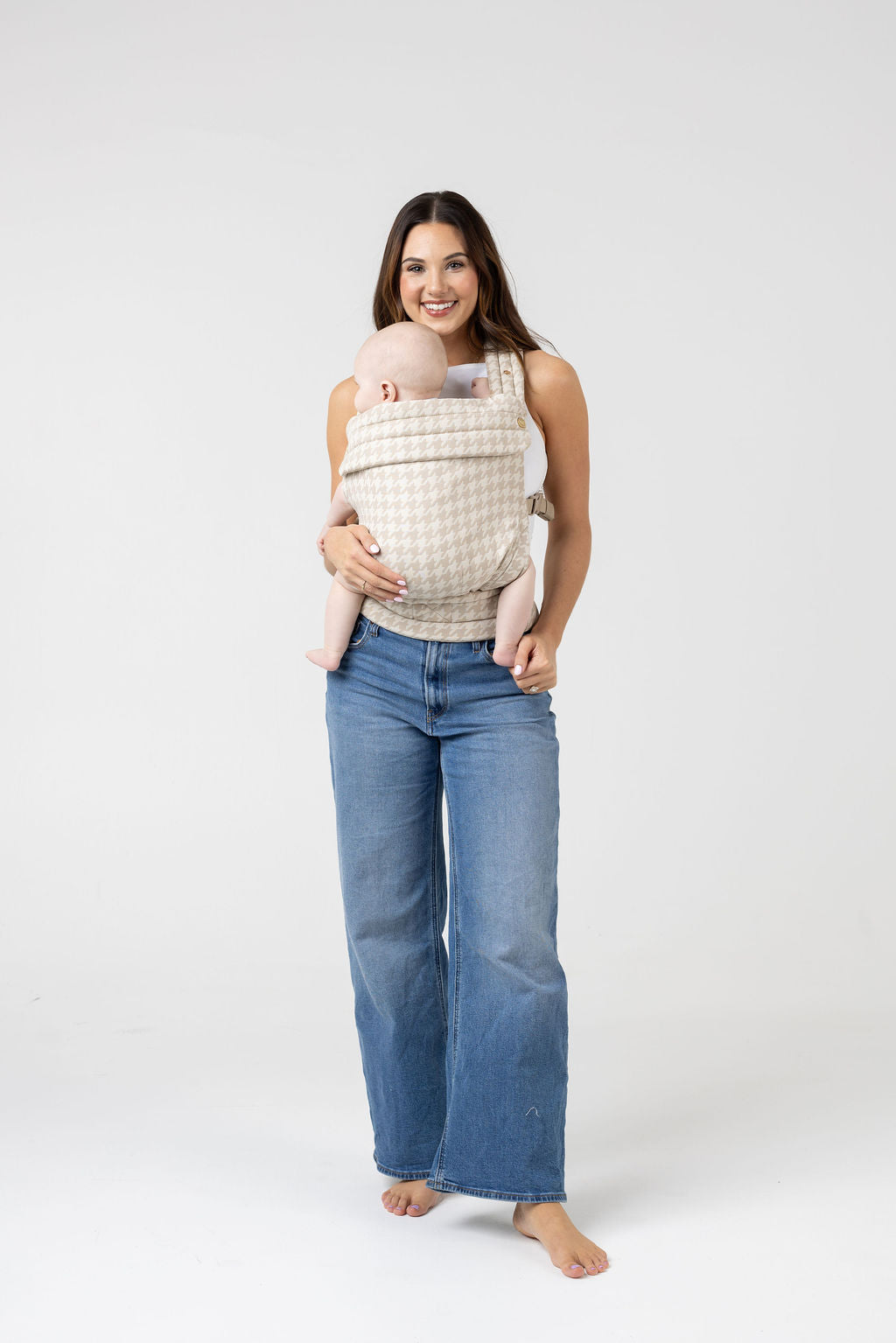 Full-length photo of a woman with long brown hair with highlights and tanned skin wearing a white tank top and the Mabē Monarch Buckle Carrier in Houndstooth, which is cream and beige. Inside is a light-skinned baby. She wears medium wash wide leg jeans and faces the viewer with a big smile. The baby is looking to the left of the frame. His legs are bare and the woman is barefoot.