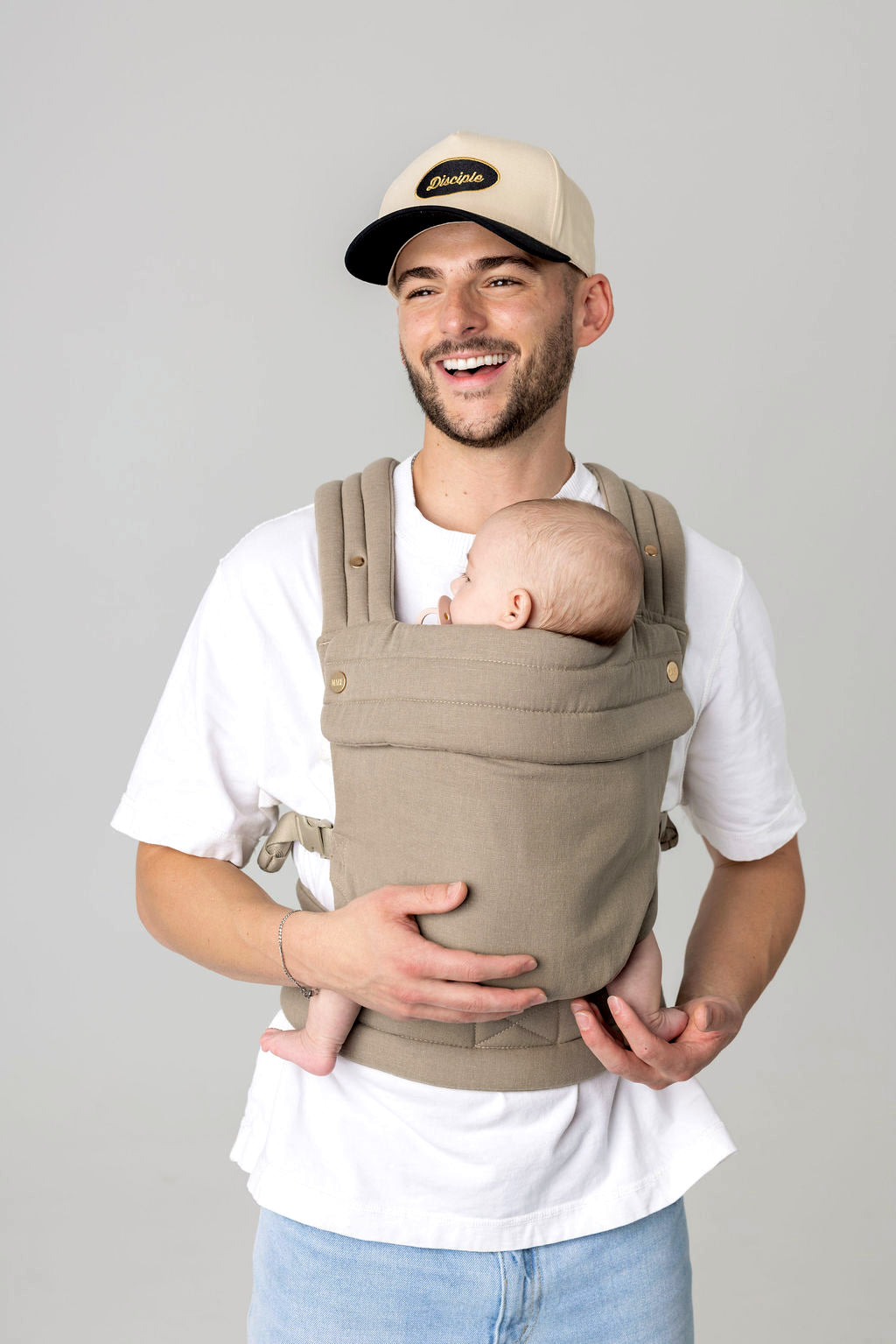 Waist-to-above-head photo of a white man with dark facial hair and a light-colored hat with a dark brim carrying a baby in a greenish-gray carrier against a light background. He is wearing a white t-shirt and light wash jeans which are only just visible.