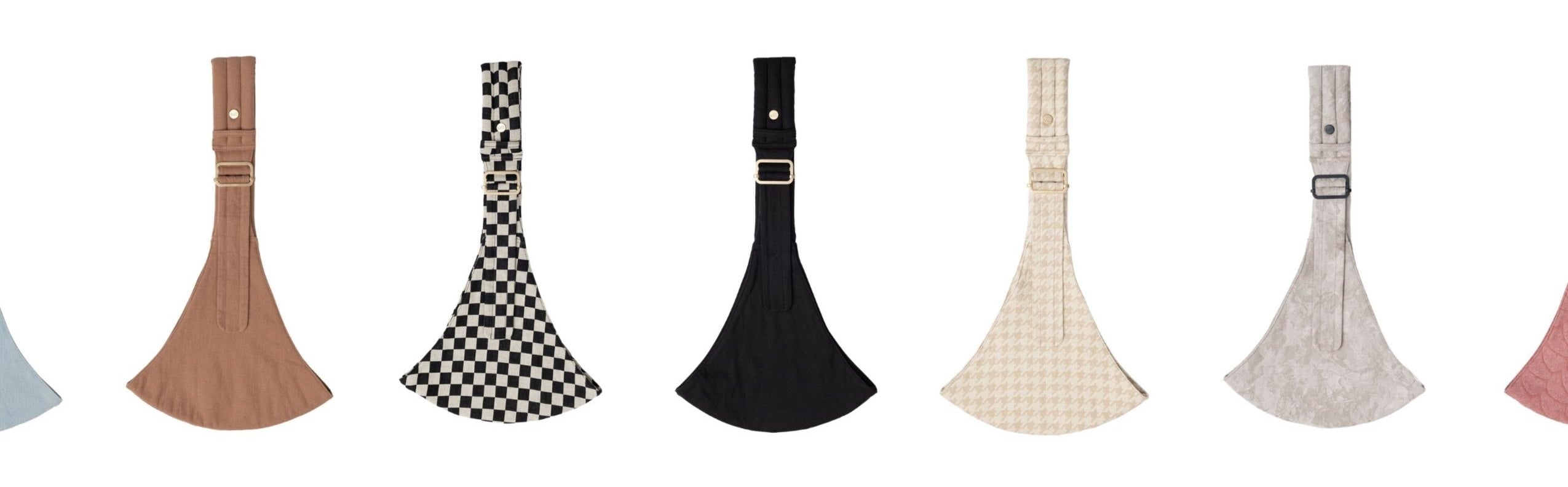 A row of seven Mabē toddler slings is displayed against a white background, each shown laid flat with its strap and buckle visible at the top. The slings come in different colors and patterns, including mocha, black‑and‑white checkered, solid black, cream houndstooth, light gray camo, and soft pink with butterfly wings. Their flared, curved shapes and textured fabrics are evenly spaced in a clean, minimal arrangement.