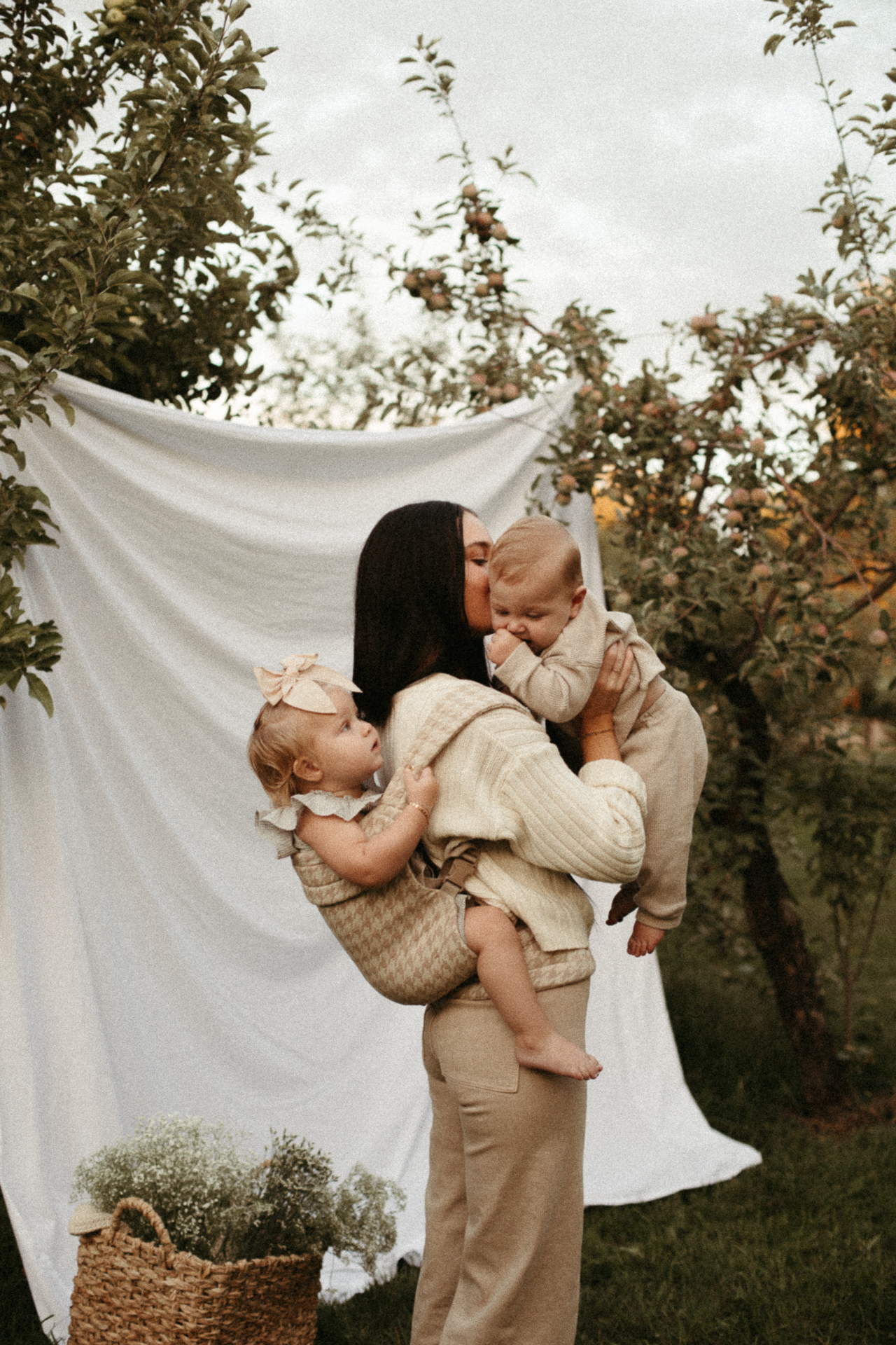A woman with very dark straight hair is facing to the right of the frame, holding a big baby up to her face and a toddler girl on her back in the cream and beige Mabē Monarch Buckle Carrier in Houndstooth pattern. They are all dressed in shades of cream and beige and are standing in front of a white backdrop strung between two fruit trees in an orchard in the background. A basket of bunches of tiny flowers or herbs is on the ground behind them.