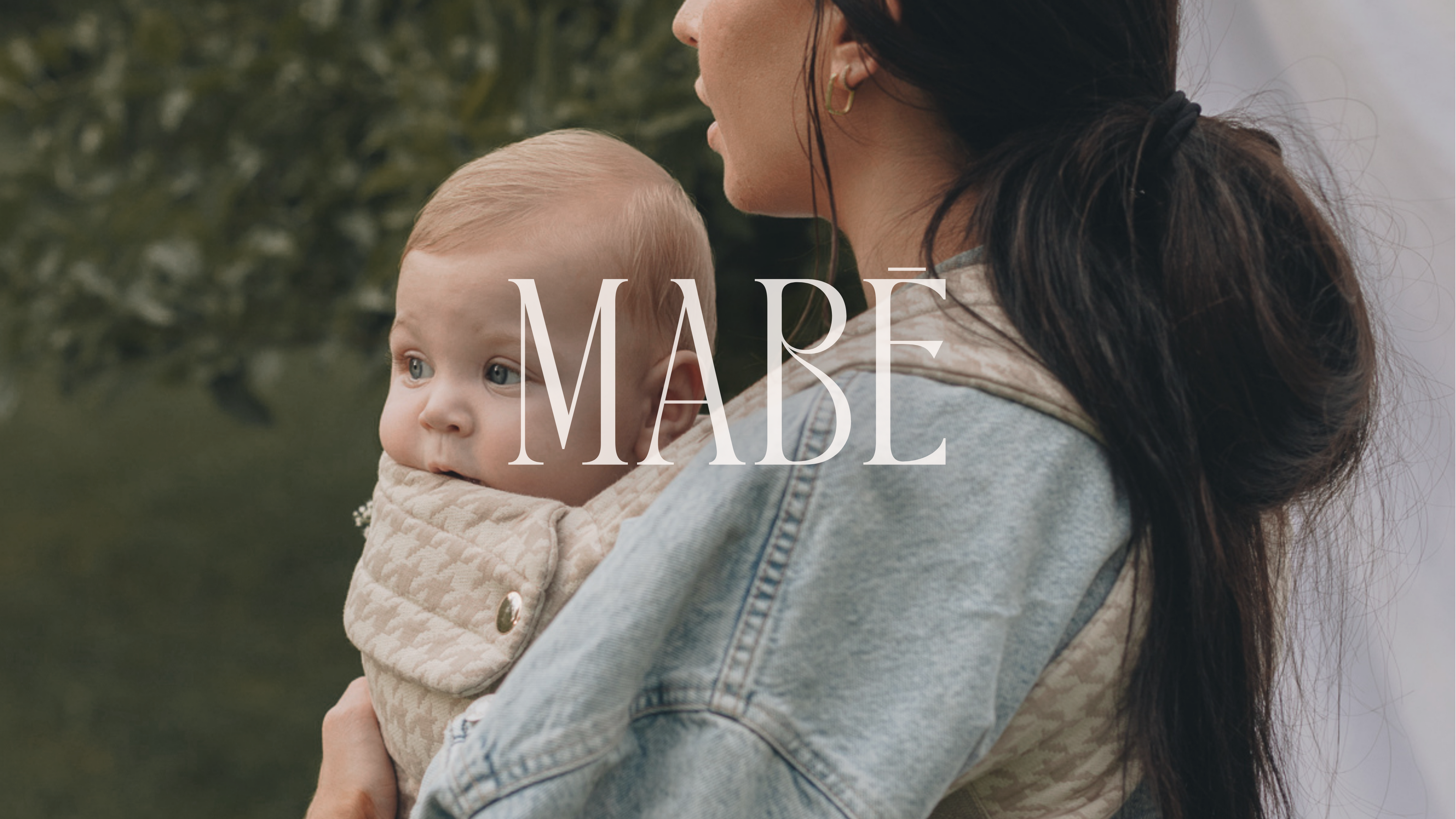 A cropped photo of a woman facing the left of the frame, with tanned skin and long straight dark hair pulled back in a loose loop. She is shown from the nose down and shoulder up, and the main focus is on a light-haired, light-skinned baby in the Mabē Monarch Buckle Carrier in Houndstooth, which is beige and cream and houndstooth-patterned. The baby is faced in toward the woman but he looks to the left of the frame. The woman is wearing a light wash denim jacket.