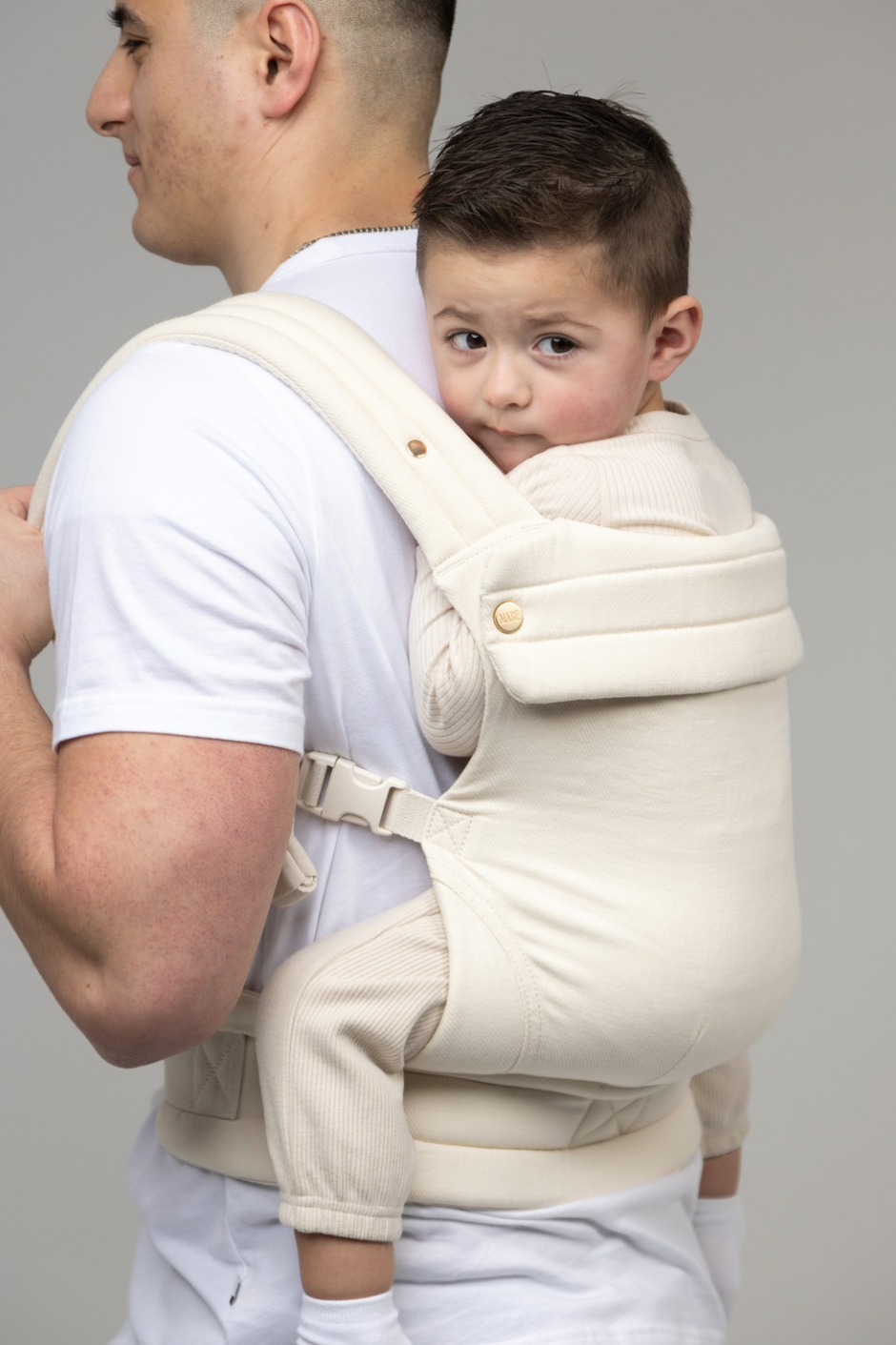 A cropped photo of a man with dark buzzed hair shown from the ears down. He faces away from the viewer and wears a white t-shirt and a toddler in the Mabē Monarch Buckle Carrier in Ivory, which is a solid warm off-white color. The toddler has dark brown hair and looks with a slightly skeptical face to the right of the frame. He is wearing cream colored coordinates.